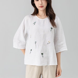 May include: A white linen top with three-quarter sleeves and a round neckline. The top is adorned with delicate embroidered floral designs in green and yellow. Paired with beige linen pants, creating a casual, comfortable outfit.