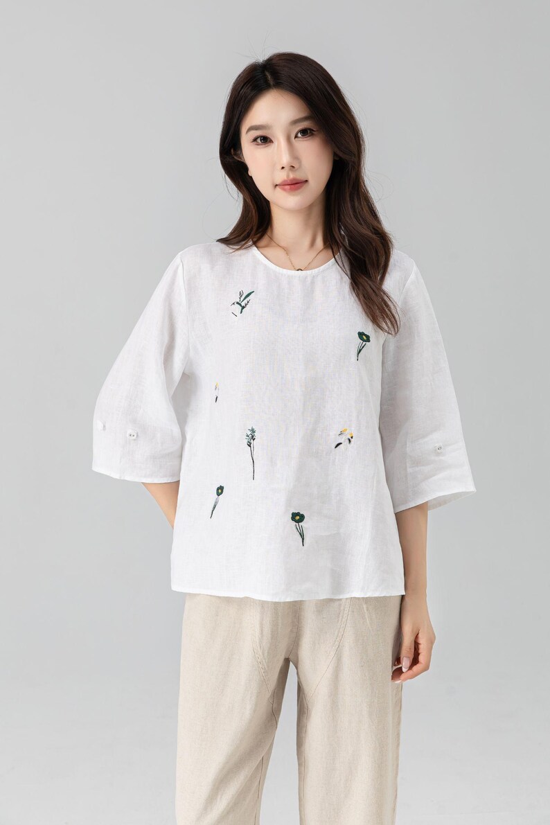 May include: A white linen top with three-quarter sleeves and a round neckline. The top is adorned with delicate embroidered floral designs in green and yellow. Paired with beige linen pants, creating a casual, comfortable outfit.
