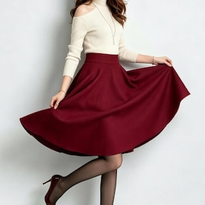May include: A cream-colored long-sleeve top with cold-shoulder cutouts and a high neckline paired with a solid deep red midi-length skirt that features a flared, A-line silhouette and a high waistband, worn with sheer black tights and dark red heels.