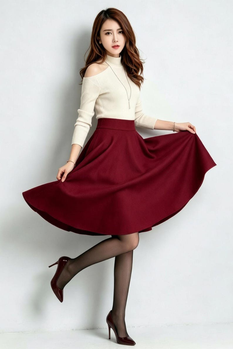 May include: A cream-colored long-sleeve top with cold-shoulder cutouts and a high neckline paired with a solid deep red midi-length skirt that features a flared, A-line silhouette and a high waistband, worn with sheer black tights and dark red heels.