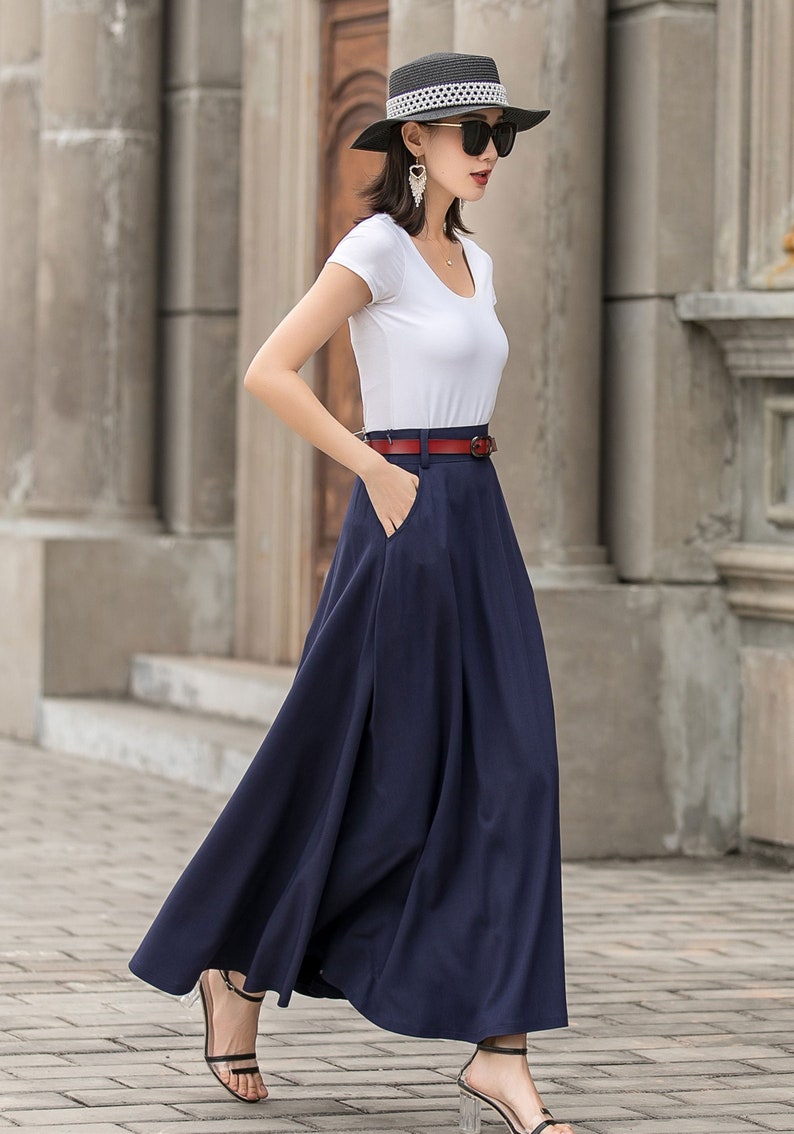 May include: A woman wearing a white scoop-neck t-shirt, a navy blue maxi skirt, and a red belt. She is also wearing a black hat, sunglasses, and sandals. The skirt has pockets.