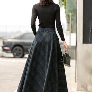 May include: A woman wearing a black turtleneck sweater and a black hat, walking down a pavement. She is wearing a long, green and black plaid skirt.