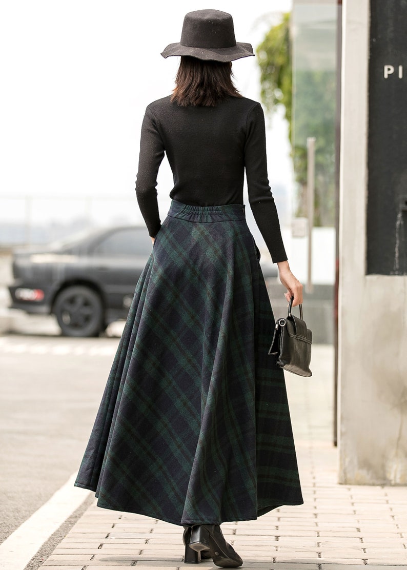 May include: A woman wearing a black turtleneck sweater and a black hat, walking down a pavement. She is wearing a long, green and black plaid skirt.