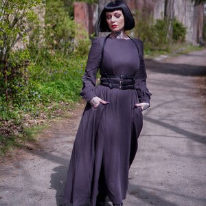 May include: A woman wearing a long, dark grey dress with a black leather harness belt. She has black hair and is standing on a path in a park.