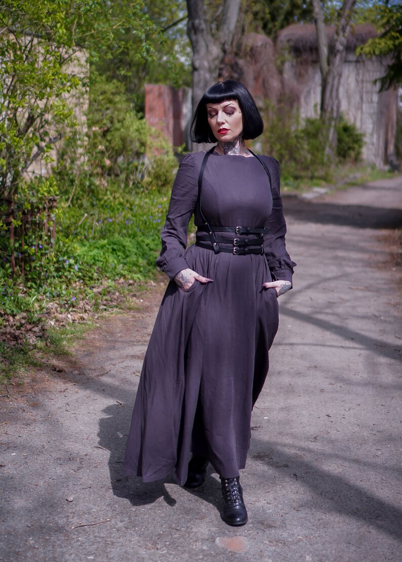May include: A woman wearing a long, dark grey dress with a black leather harness belt. She has black hair and is standing on a path in a park.