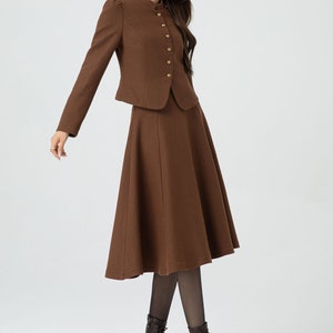 May include: A brown skirt suit with a fitted jacket and a flared skirt. The jacket has a mandarin collar and gold buttons. The skirt is knee-length and has a slight A-line shape.