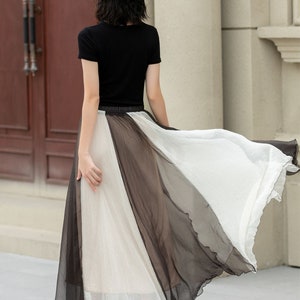 May include: A woman wearing a black short-sleeved shirt and a white hat, walking in a black and white sheer maxi skirt.