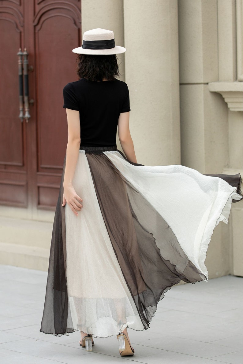 May include: A woman wearing a black short-sleeved shirt and a white hat, walking in a black and white sheer maxi skirt.