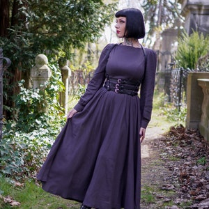May include: A woman wearing a long, dark purple dress with a black leather corset and black boots. She is standing in a cemetery with trees and tombstones in the background.