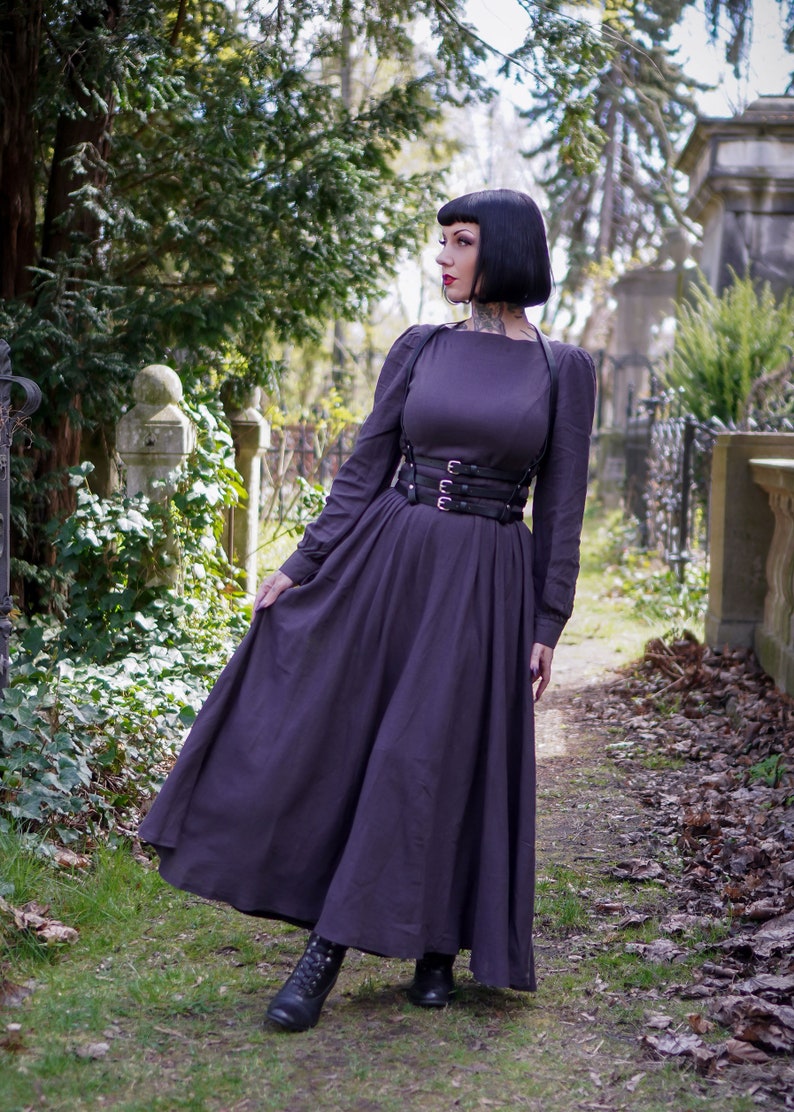 May include: A woman wearing a long, dark purple dress with a black leather corset and black boots. She is standing in a cemetery with trees and tombstones in the background.