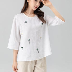 May include: White linen top with embroidered floral details. The top has a round neckline and three-quarter sleeves. The floral embroidery features green and yellow accents. Paired with beige linen pants.