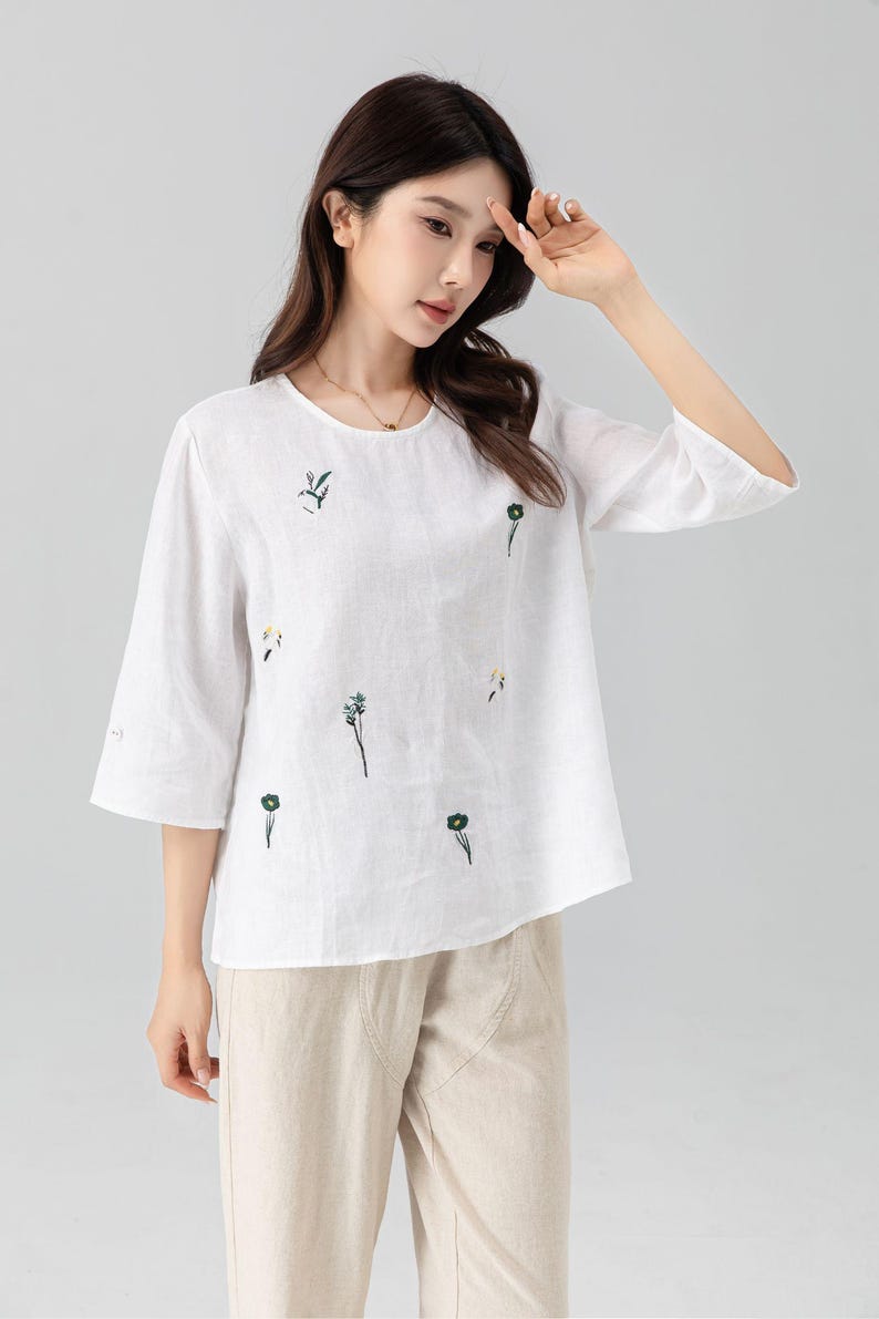 May include: White linen top with embroidered floral details. The top has a round neckline and three-quarter sleeves. The floral embroidery features green and yellow accents. Paired with beige linen pants.