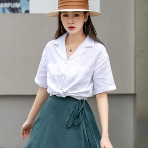 May include: A woman wearing a white linen shirt with short sleeves tied at the waist, a dark green wrap skirt, and a brown and white wide-brimmed hat.