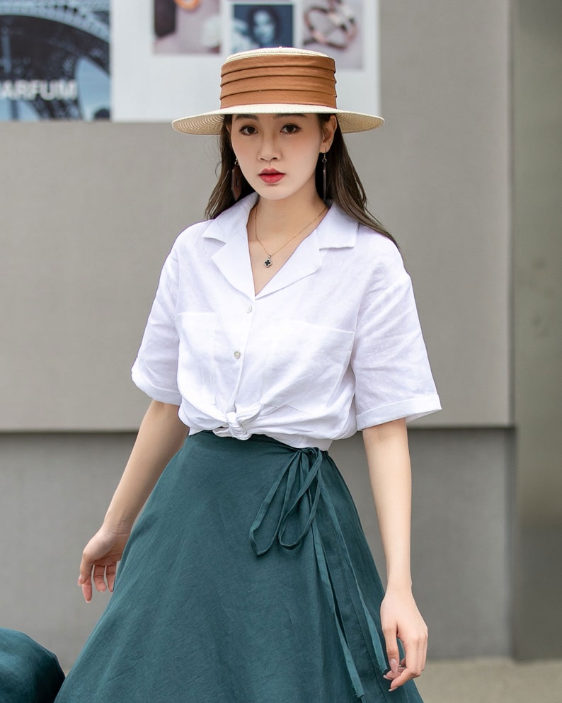 May include: A woman wearing a white linen shirt with short sleeves tied at the waist, a dark green wrap skirt, and a brown and white wide-brimmed hat.