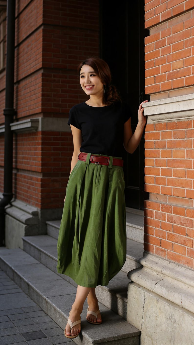 May include: A woman wearing a black t-shirt, a green skirt, and a red belt. The skirt is pleated and falls to the mid-calf. She is wearing beige sandals. The background features a brick building.