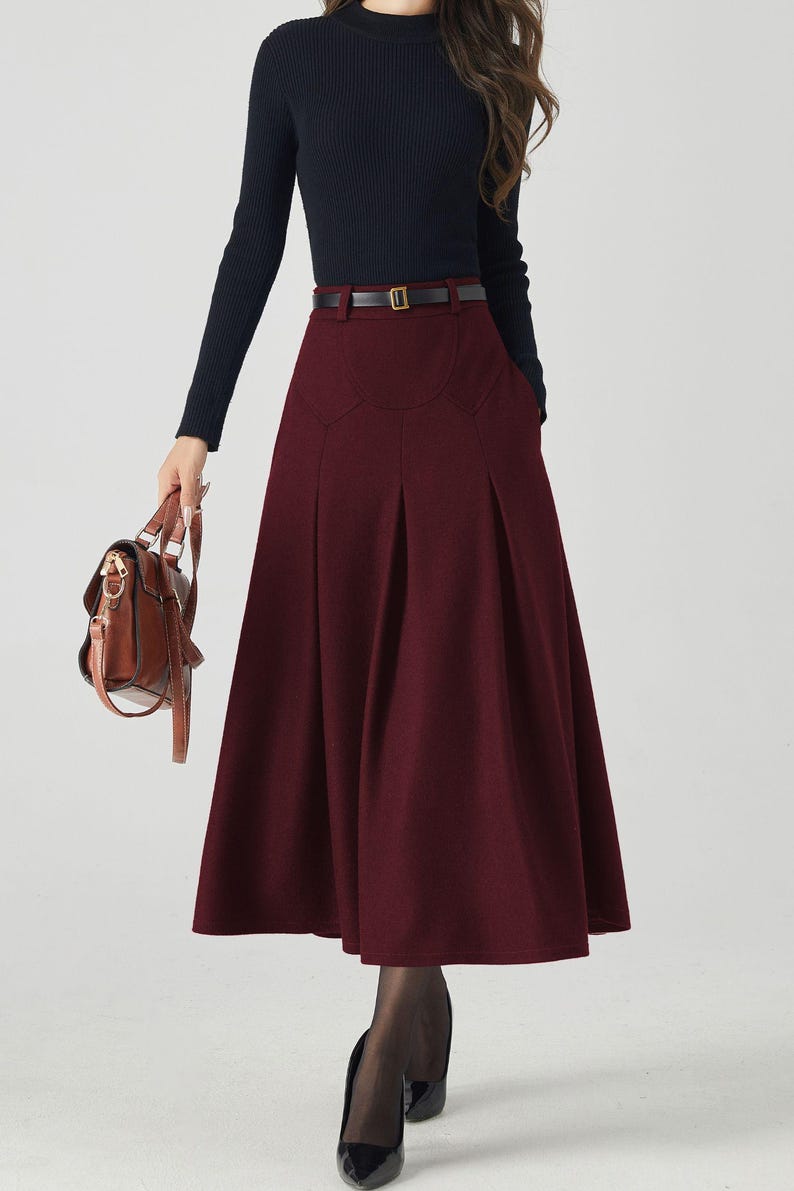 May include: A burgundy midi skirt with pleats and a black belt. The skirt is paired with a long-sleeved navy blue top and a brown leather handbag. The outfit is completed with black tights and heels, creating a classic, elegant look.
