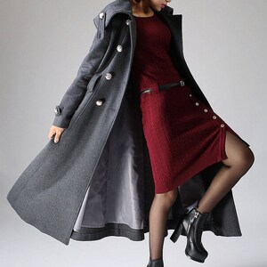May include: A long gray wool coat with a high collar and large buttons. The coat is worn over a red knit dress and black tights. The person is wearing black heeled boots.