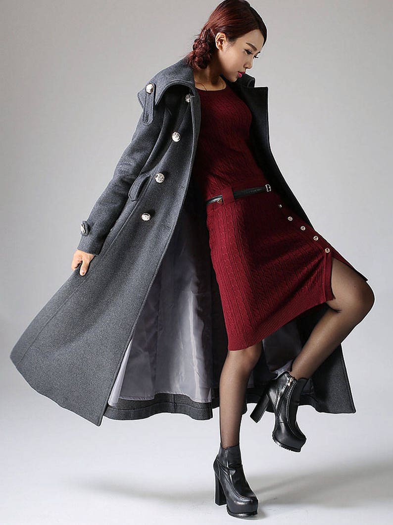 May include: A long gray wool coat with a high collar and large buttons. The coat is worn over a red knit dress and black tights. The person is wearing black heeled boots.