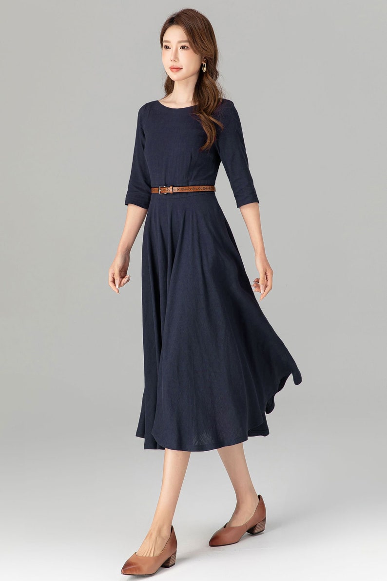 May include: A navy blue midi dress with a brown belt. The dress has a round neckline and 3/4 sleeves. The model is wearing brown heeled shoes.