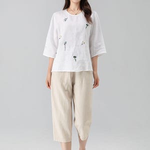 May include: White linen top with embroidered floral details and three-quarter sleeves, paired with beige capri pants. The outfit is a casual, comfortable style, suitable for warm weather. The pants are approximately knee-length.