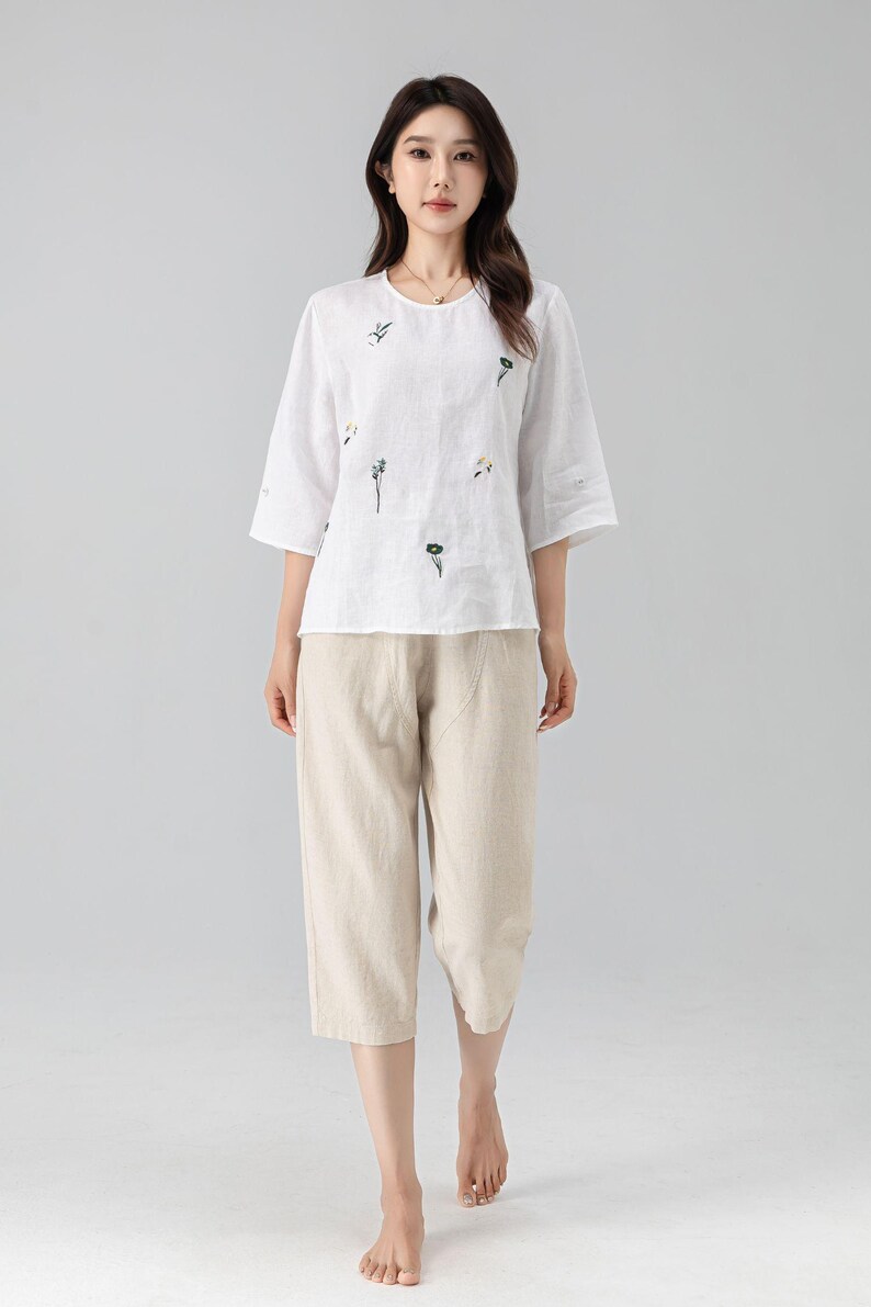 May include: White linen top with embroidered floral details and three-quarter sleeves, paired with beige capri pants. The outfit is a casual, comfortable style, suitable for warm weather. The pants are approximately knee-length.