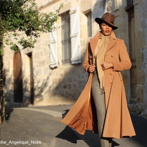 May include: A woman wearing a long camel-colored coat with a brown hat and a beige scarf. The coat has a double-breasted closure and a flared skirt. The woman is standing on a cobblestone street.