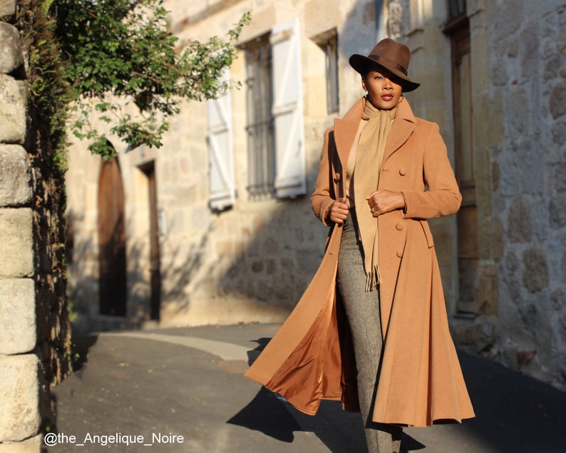 May include: A woman wearing a long camel-colored coat with a brown hat and a beige scarf. The coat has a double-breasted closure and a flared skirt. The woman is standing on a cobblestone street.