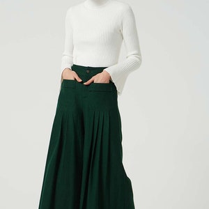 May include: A woman wearing a white turtleneck sweater and dark green wide-leg trousers with pleats and large pockets.