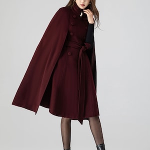 Pu&ograve; includere: Un cappotto a mantella color bordeaux con collo alto decorato da due bottoni perlati, chiusura a doppio petto con bottoni in tessuto coordinati e cintura da annodare. Il capo &egrave; indossato sopra una maglia nera a maniche lunghe.