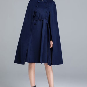 Pu&ograve; includere: Un cappotto a mantella blu navy lungo fino al ginocchio con colletto alto a volant, tre bottoni bianchi effetto perla, chiusura doppiopetto con bottoni blu navy abbinati e cintura da annodare. Abbinato a stivaletti neri.