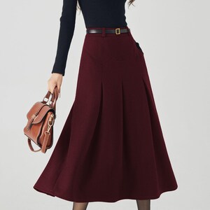 May include: A woman's outfit featuring a black long-sleeved top and a burgundy midi skirt with a belt. The outfit is accessorized with a brown handbag and black high heels. The skirt has a pleated design.