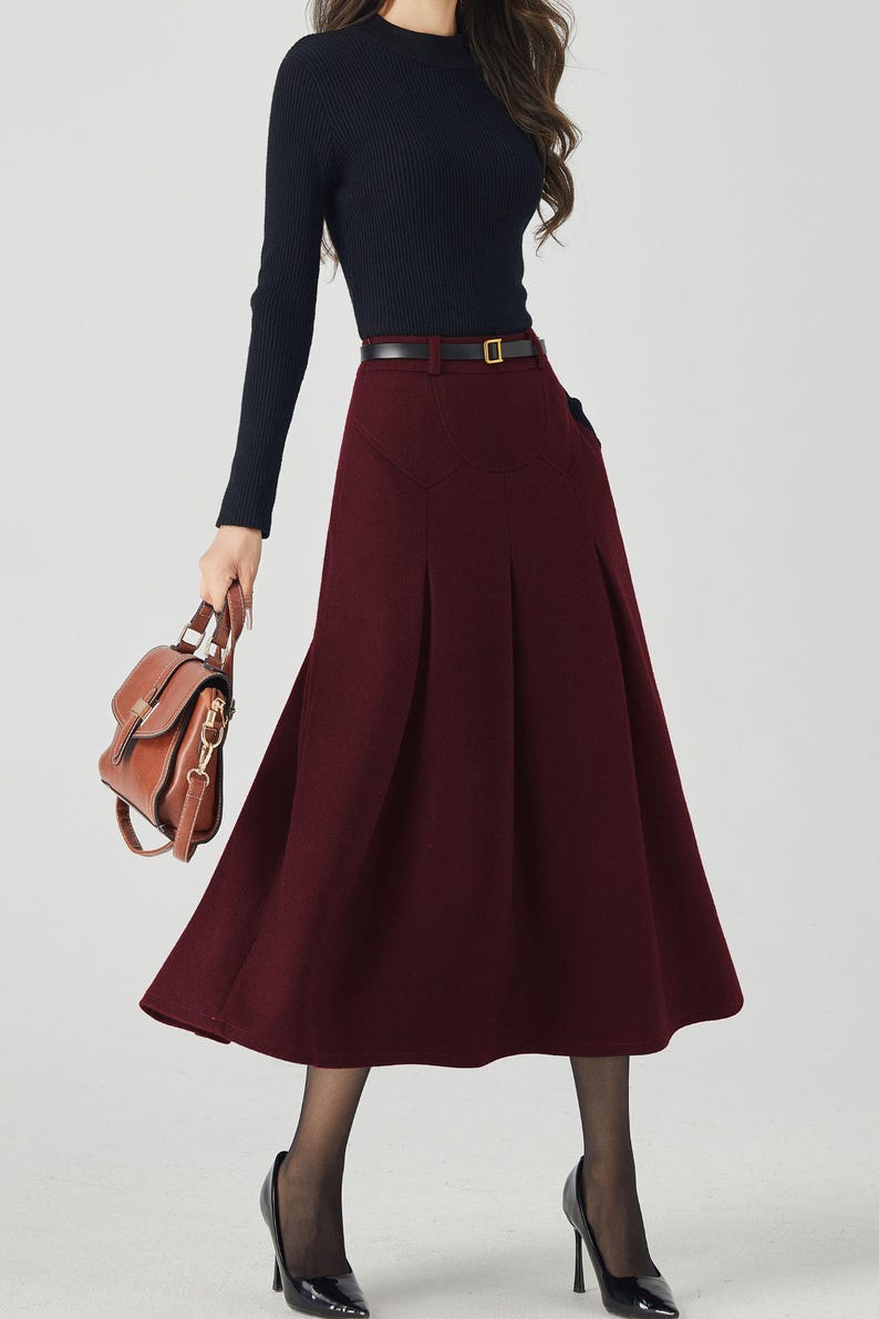 May include: A woman's outfit featuring a black long-sleeved top and a burgundy midi skirt with a belt. The outfit is accessorized with a brown handbag and black high heels. The skirt has a pleated design.