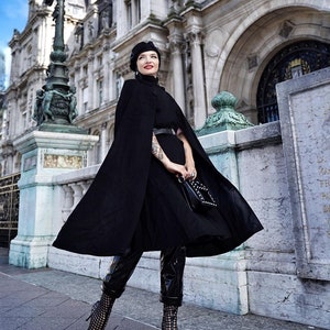 May include: A woman wearing a black cape, black pants, and black boots with a black purse in front of a building.