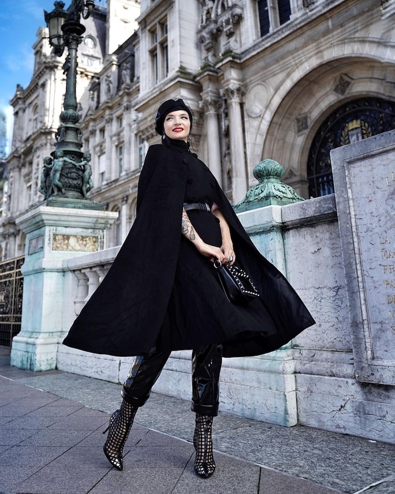 May include: A woman wearing a black cape, black pants, and black boots with a black purse in front of a building.