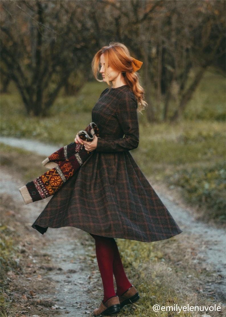 Pu&ograve; includere: Una donna indossa un abito a quadri marroni e neri con gonna lunga e maniche lunghe. Tiene in mano un maglione di lana colorato. Indossa collant rossi e scarpe nere. La donna &egrave; in piedi su un sentiero in un bosco.