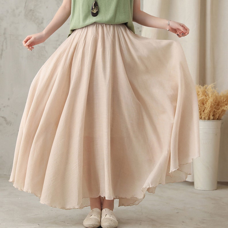 May include: A flowing, beige maxi skirt with a wide, gathered waistband. The skirt is made of a lightweight fabric that drapes gracefully. The hemline has a subtle, scalloped edge. The skirt is part of a casual outfit.