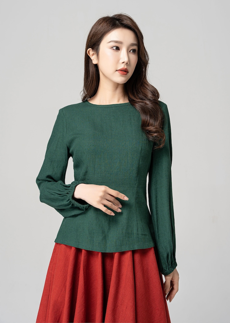 May include: A green long-sleeved blouse with a round neckline and gathered cuffs. The blouse is tucked into a red skirt.