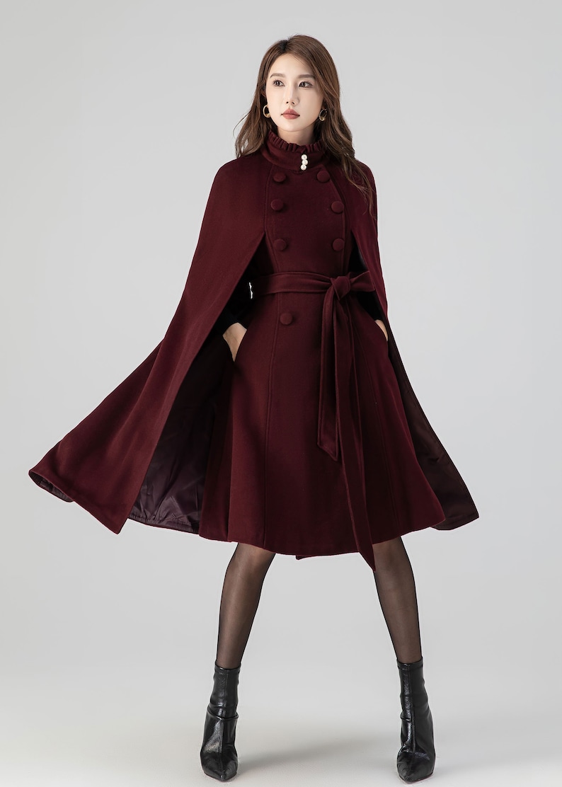 Pu&ograve; includere: Un cappotto a mantella in lana color bordeaux con colletto alto a volant, tre bottoni effetto perla, chiusura doppiopetto con bottoni in tessuto coordinati e cintura in vita. Il capo presenta tasche laterali e una linea svasata.