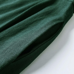 Puede incluir: Una prenda de color verde bosque oscuro que presenta un bolsillo lateral visible. La tela muestra una textura tejida distintiva con sutiles pliegues verticales y frunces que se extienden desde la abertura del bolsillo, creando un aspecto suave.