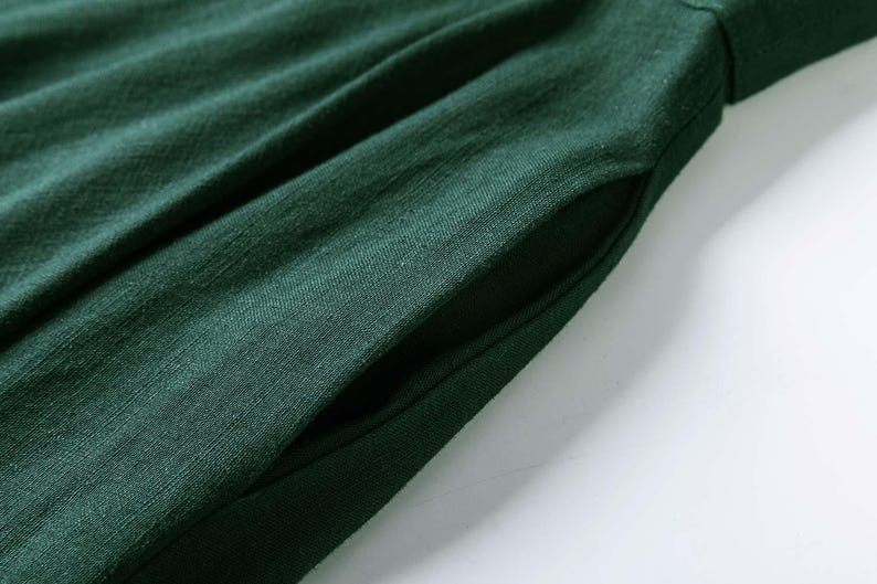 Puede incluir: Una prenda de color verde bosque oscuro que presenta un bolsillo lateral visible. La tela muestra una textura tejida distintiva con sutiles pliegues verticales y frunces que se extienden desde la abertura del bolsillo, creando un aspecto suave.