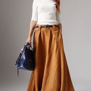 May include: A woman wearing a white top and a long, brown, linen skirt with a black belt. She is holding a blue handbag with a patterned scarf.