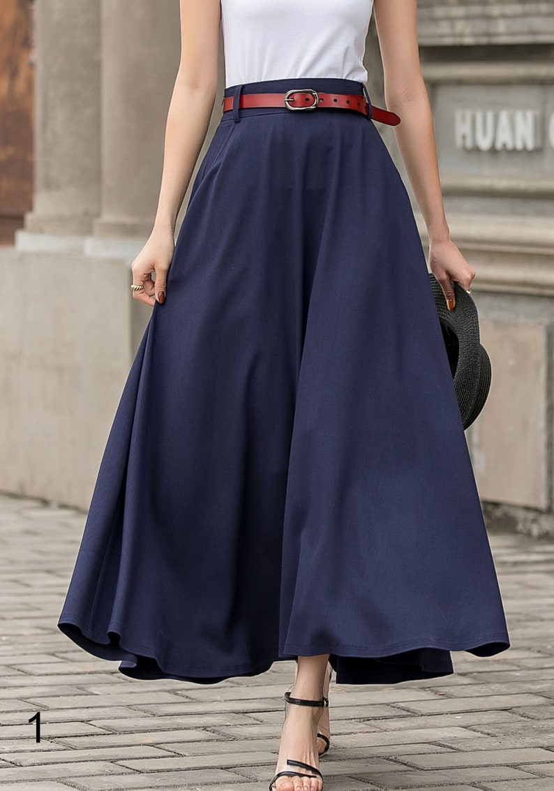 May include: A navy blue, full-length skirt with a wide, flowing silhouette. The skirt is paired with a red belt and a white top. The outfit is completed with black sandals and a black hat.