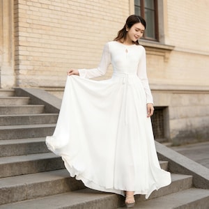 Puede incluir: Un vestido largo y blanco, con mangas largas y escote de ojo de cerradura. El vestido tiene una cintura ce&ntilde;ida con un lazo y una falda larga hasta el suelo. La mujer est&aacute; de pie en unos escalones de piedra.