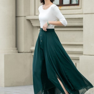 May include: A woman wearing a white top and a long, dark green skirt with a button closure and a slit in the front. She is wearing white heeled sandals.