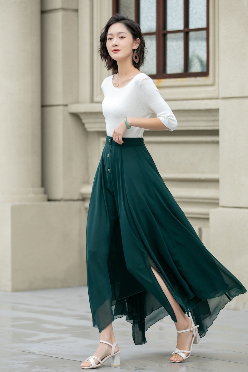 May include: A woman wearing a white top and a long, dark green skirt with a button closure and a slit in the front. She is wearing white heeled sandals.