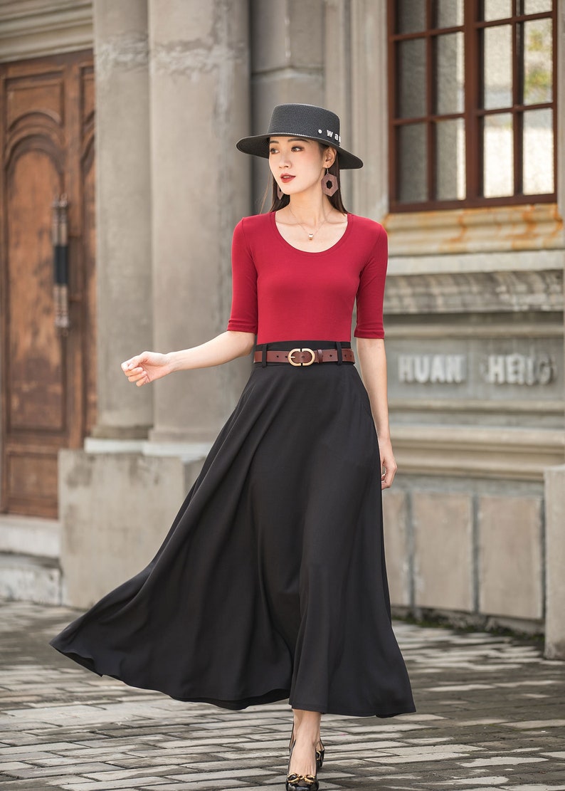 May include: A woman wearing a red scoop-neck top with elbow-length sleeves and a long black skirt. She is wearing a black hat and a brown belt with a gold buckle. The outfit is suitable for a formal event.