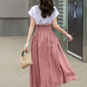 May include: A woman wearing a white short-sleeved shirt and a long, dusty rose-colored skirt. She is carrying a small, woven straw bag and walking away from the camera.