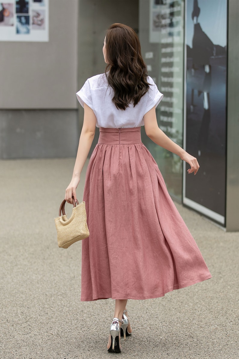 May include: A woman wearing a white short-sleeved shirt and a long, dusty rose-colored skirt. She is carrying a small, woven straw bag and walking away from the camera.