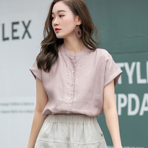 May include: A woman wearing a light pink linen shirt with a button-up front and short sleeves. She is also wearing a light beige linen skirt with a drawstring waist.