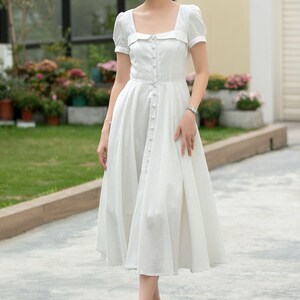 Pode incluir: Um vestido midi branco com decote quadrado, mangas curtas e fecho de botões na frente. O vestido tem uma saia ligeiramente rodada e é feito de um tecido leve.